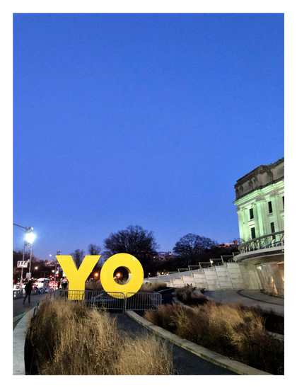 The front plaza of the Brooklyn Museum, under a clear blue evening sky. At right is the museum’s facade. At left, giant yellow aluminum letters spelling out “YO” sit behind guardrails next to bushes and a busy street.