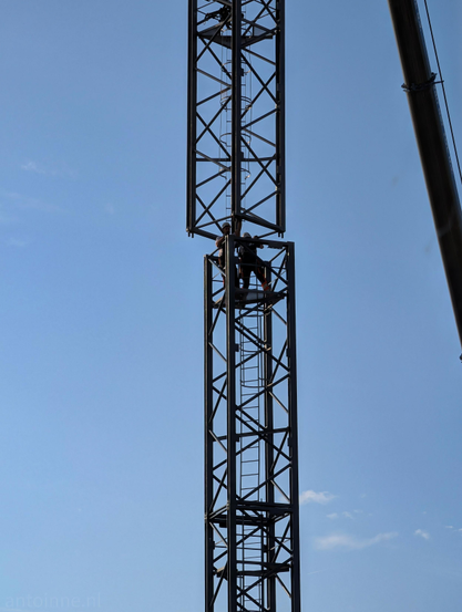 Workers constructing a dark metal crane structure against a clear blue sky. Inside the structure, vertical ladders and safety hoops run up the core.

Two construction workers equipped with hard hats and work boots are positioned at the junction where the two mast sections connect. They are guiding the upper section into place.

The background features a bright, clear blue sky with light, faint wisps of clouds near the bottom right. 