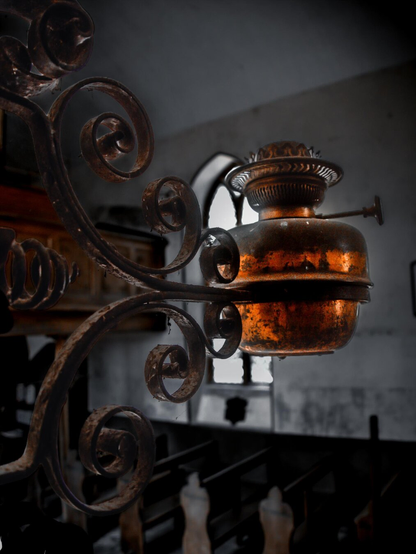 A weathered, vintage kerosene lamp burner made of aged copper or brass with an amber patina. The metallic fixture is held by thick wrought-iron details covered in light dust. In the background, rows of wooden church pews are illuminated by the light coming through an arched window.