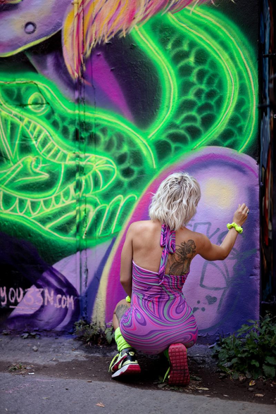 a photo of Psyqu33n, she is signing her wall. And she is wearing the same vivid colors she used on it: light purple and green.