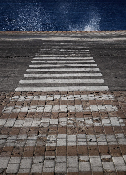 A photo of a zebra crossing over a paved walkway leading towards deep blue water. A plume of white water can be seen where the crossing ends.