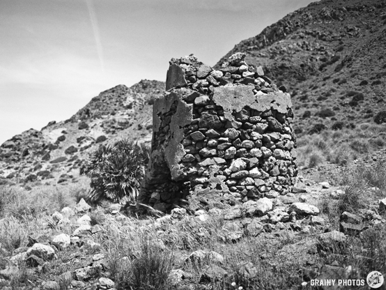 A black and white image of a weathered stone structure surrounded by rocky terrain and sparse vegetation, set against a backdrop of rolling hills under a clear sky.