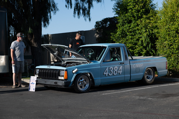 A light blue truck, lowered on modern wheels, with its hood open showing a modern engine. An exhaust pipe sticks out forward of the front wheel. The racing number 4384 is painted on its side. People are standing around it.