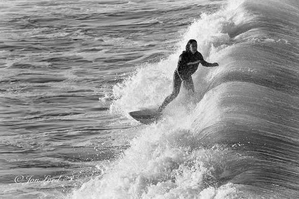 This is a black and white water sports photo in landscape format of a female teen surfing shortly after sunrise. Hermosa Beach, California (2013).

Stretching from the right edge of the photo to a little less than the half way across point is a fierce braking Pacific wave. The wave which is now a little less than two metre high, is moving from right to left, viewed from two metres above and a few metres away from the closest point. The wave has multiple streaks or lines extending at right angles to the wave, from the crest and out to sea. Ahead of the breaking wave is our young female surfer. She is positioned slightly to the right of centre in the maelstrom of white foaming surf and is in motion diagonally across the photo from the centre towards the lower left corner. Our surfer is viewed from the front, with her standing on the board sideways (standing on the board and facing to the left). She is viewed from above at five metres distant and with her occupying half the height of the photo. She is wearing a wetsuit with a the name 'Redondo' in large white lettering on the front. She has long dark and wet hair. Her left arm is held above the breaking wave as she maintains her balance, with her left leg inside the wave and her right arm is held downwards. Ahead of our surfer and the wave, is an area of moderately smooth water with lines of white foam running at right angles to the surf marking the passage of previous waves. 

This was the morning surfing rush hour, just after sunrise, when many teens would dash to the beach for an hour or so of surfing before reluctantly ambling off to school. Oh, and there were one or two adults in the surf as well. 