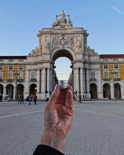 A forced-perspective photo features the historic Rua Augusta Arch (Arco da Rua Augusta) in Lisbon, Portugal, aligned to look like a giant lightbulb base held in a person's hand.