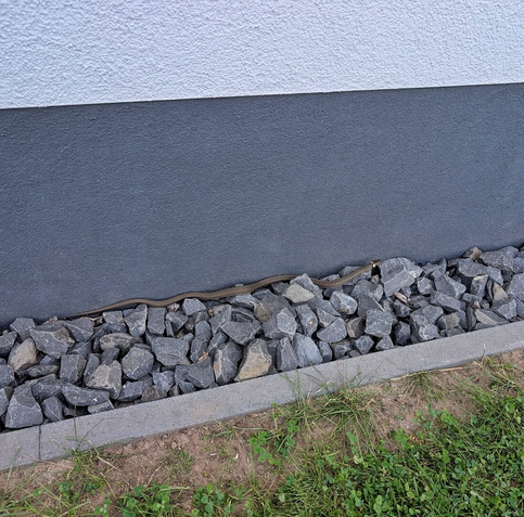A grass snake slithers across a bed of uniform grey decorative stones beside a smooth dark grey wall. Low-growing green grass and clover fill the foreground as the snake moves along the edge of the stone border.