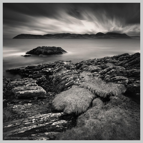 A photograph of the view across the Firth of Clyde towards the Isle of Arran from the Kintyre peninsula, presented in monochrome. The calm, smooth water fills the foreground and middle distance, reflecting the soft effect of a long exposure, while Arran appears on the horizon as a dark, rugged silhouette beneath a heavy, brooding sky. The contrast between the stillness of the water and the dramatic cloud-filled sky gives the scene a stark, atmospheric quality.