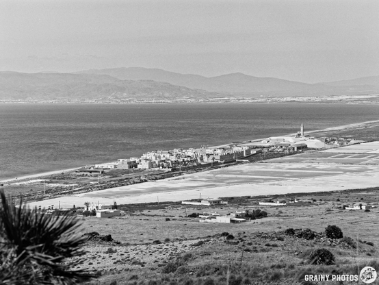 A panoramic view of Las Salinas and a coastal village with buildings along the shore, set against the backdrop of distant mountains. The scene is captured in black and white, emphasizing the natural landscape and the serene waters of the sea.