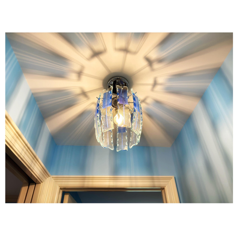 Photo looking up at a lightbulb suspended from a ceiling and surrounded by nine vertical glass panels. These are slightly curved with irregular edges, and each has a narrow bluish vertical strip flanked by two bluish rectangles painted on the otherwise clear glass. The bulb’s light shining through the panels casts a striking display of blue stripes on the ceiling, radiating out from a central hub and continuing down the walls of the room. The upper parts of doorways can also be seen at the bottom of the picture.