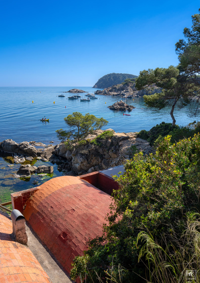 Color photo of a rocky shoreline on a blue sea with many rocks and small islands sticking up from the water and people paddling a small boat and the tiled, barrel-vaulted roofs of fishermen’s houses in the foreground below.