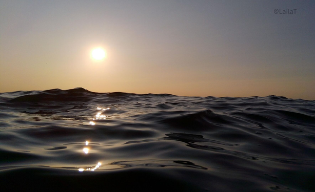 Meerwasser mit Wellen kurz über der Oberfläche fotografiert zur tiefstehenden Sonne hin, die auf dem Wasser funkelt und den Himmel orange färbt.