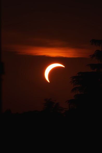 This photograph is of a partial solar eclipse setting against a deep, dark red sky (really low f stop). There is a bright crescent of light which shines brightly in the centre, partially covered by a horizontal band of orangey/red clouds above it. On the right there are dark silhouetted trees which frame it.
