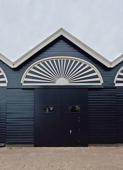 A photo of a part of a dark wooden building with a large  door and a large white fan-shaped decorative element above it. The building has gabled roofs with a white trim.