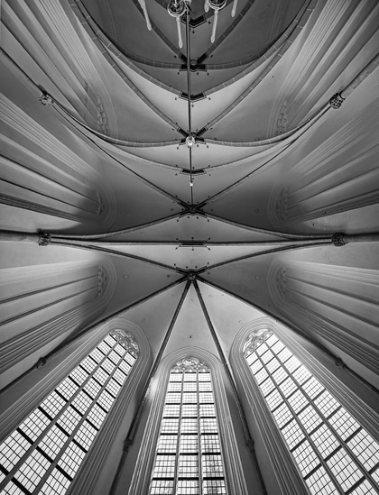 Black-and-white symmetrical view straight up at a Gothic church ceiling, showing ribbed vaulting radiating outward in a fan pattern from a central line of bosses, with tall pointed lancet windows visible at the bottom of the frame.