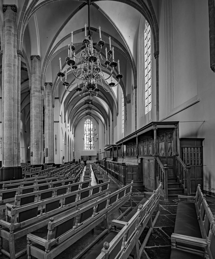 Black-and-white interior view of a Gothic church nave, looking down rows of empty wooden pews toward a distant stained-glass window, with ribbed vaulted ceiling arches overhead and an ornate wrought-iron candle chandelier hanging in the foreground; carved wooden choir stalls line the right side.


Next time, try this in Cowork