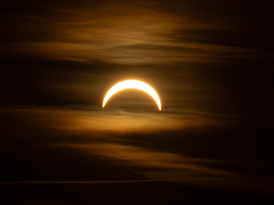 Ein orangener mit leichten Wolken durchsetzter Himmel. im Zentrum eine schmale Sichel der Sonnenscheibe, die Sichel ist senkrecht nach unten geöffnet