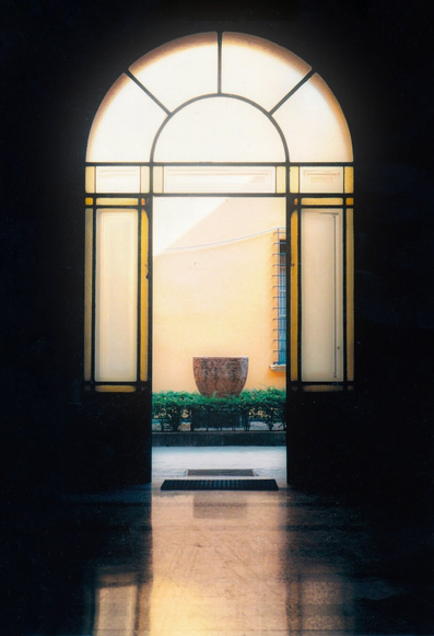 A view down a corridor to an open doorway onto a courtyard. In the courtyard there's a low green shrubbery border and a large terracotta vase. The doorway is set in a glass arch, clear in the upper arch with yellow stained glass panels down the sides. The bottom third of the photo is the light reflecting off the shiny pavement, while the top and sides are in shadow.