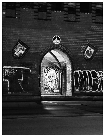 Schwarz-Weiß-Foto im Hochformat mit weißem Rand. Zu sehen ist ein Teil der Oberbaumbrücke in Berlin bei Nacht. An den dunklen Backsteinen sind Graffiti, dazwischen ein beleuchteter Torbogen, durch den man ein weiteres Graffito sieht - einen Vampirkopf. Er scheint aus dem Torbogen zu schauen. 
