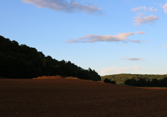 Photograph of a rural landscape at the end of the day, showing a plowed field of brown soil, now in shadow except for a few mounds of earth in the middle, illuminated by the setting sun. Behind, at the edge of the field, a dark green hill rises to the left. A little further behind and to the right, another green hill in the sun. The sky is still a deep blue, with a few small white clouds reflecting the light of the setting sun.

Photographie d'un paysage rural en fin de journée, montrant un champ retourné de terre brune, maintenant dans l'ombre, sauf quelques tas de terre en plein milieu, éclairés par le soleil couchant. Derrière en bordure du champ, une colline vert sombre du côté gauche. Un peu plus loin derrière et à droite, une colline verte au soleil. Le ciel est encore d'un bleu intense, avec quelques petits nuages blancs réfléchissant la lumière du soleil couchant.