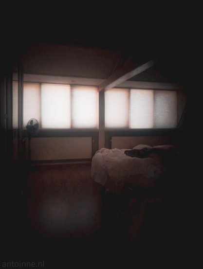 A dimly lit loft bedroom with an angled ceiling and a wooden support beam. Long, white heating radiators run along the wall directly under the windows.

The scene has a moody, dreamlike quality, illuminated almost entirely by soft daylight filtering through translucent window shades.

Located on the right side of the frame, an unmade bed features rumpled white sheets and pillows, suggesting someone has recently woken up or left the room.