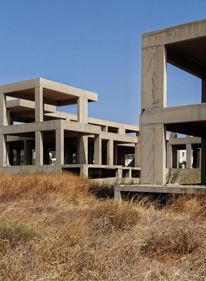 A photo of unfinished buildings, showing raw concrete structures with multiple levels and open spaces. There is dry yellow grass at the bottom and clear blue sky at the top.