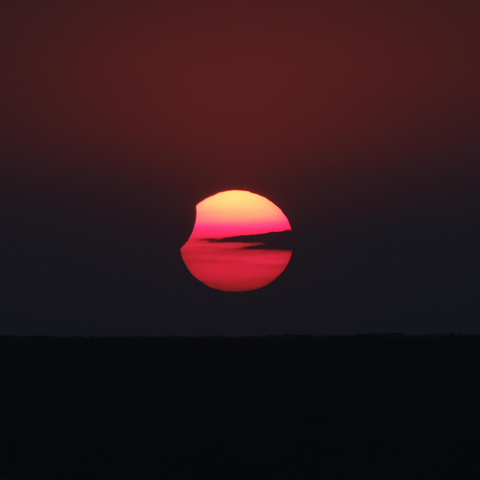 Soleil couchant avec un petit bout en haut à gauche grignoté par la Lune. Il offre un dégradé du rouge au jaune de bas en haut. Un nuage barre le soleil en son centre