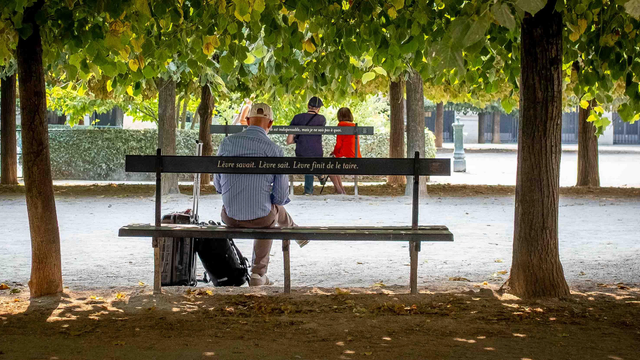 Home assegut en un banc, que mira cap a un altre banc enfront seu amb dues persones assegudes.