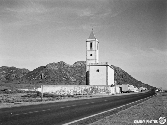 A black and white photo depicting a lone church in Las Salinas with a tower beside a road, set against a mountainous landscape under a clear sky. The scene conveys a sense of solitude and the tranquility of rural life.
