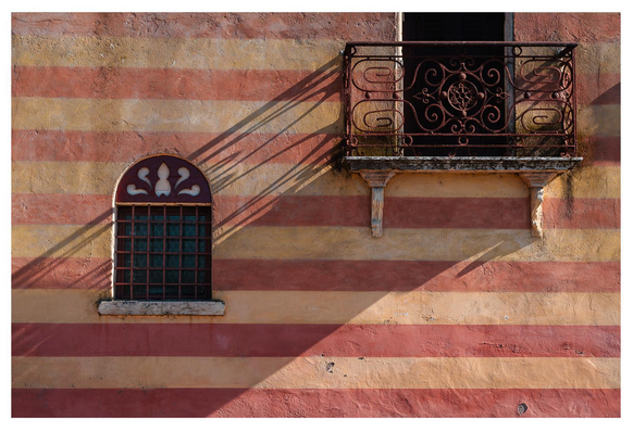 Ein rot-gelb gestreiftes Gebäude in italien, shabby chic, im Streiflicht direkt frontal aufgenommen. Ein kleines Balkonchen wirft seinen Schatten entlang der Mauer in Richtung eines kleinen Fensters.
