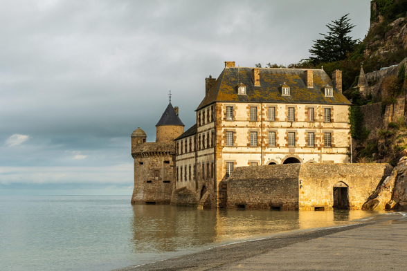 Das Bild zeigt eine beeindruckende historische Architektur, die direkt am Wasser liegt. Ein massiver Steinturm mit einem dunklen, spitzen Dach ragt links empor und scheint Teil eines größeren Gebäudekomplexes zu sein. Daran schließt sich ein helles, mehrstöckiges Gebäude mit zahlreichen Fenstern und einem charakteristischen, mit Moos bewachsenen Dach an. Die Fassade ist in einem warmen, beigefarbenen Ton gehalten und wirkt alt und geschichtsträchtig. Vor dem Hauptgebäude befindet sich eine niedrigere Steinmauer, die in das Wasser hineinragt und einen Torbogen freigibt. Der Wasserspiegel ist hoch und reicht bis an die Mauern heran, was dem Ganzen eine maritime und leicht isolierte Atmosphäre verleiht. Der Himmel ist bewölkt, aber das Licht, das durch die Wolken bricht, wirft warme Töne auf die Steine und das Gebäude, was auf einen  Sonnenaufgang hindeutet. Der Strand im Vordergrund ist mit Kies bedeckt und sanft zum Wasser hin abfallend. Die Szene strahlt eine ruhige, aber auch majestätische Stimmung aus, die an eine mittelalterliche Festung oder ein altes Kloster erinnert, das den Gezeiten trotzt.