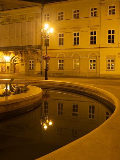 Nächtlicher Stadtplatz in warmem gelbem Laternenlicht. Im Vordergrund zieht sich der geschwungene Rand eines Brunnens durchs Bild. Das dunkle, ruhige Wasser spiegelt die beleuchtete Häuserfassade und eine mehrarmige Straßenlaterne fast vollständig. Links ist ein historisches Gebäude teilweise verhüllt; ein Blumenkorb hängt an der Laterne.