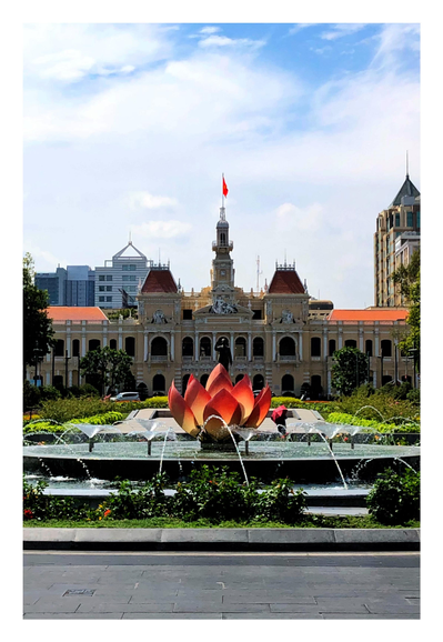 A giant lotus flower sculpture in shades of pink, yellow, and orange stands in the middle of a wide pool ringed by fountains of arcing water. In the background is a statue of Ho Chi Minh in front of a wide two-story building, with two attic towers on either side of a central clock tower flying the red flag of Vietnam. The sky overhead is overcast, with white clouds giving way to glimpses of blue.