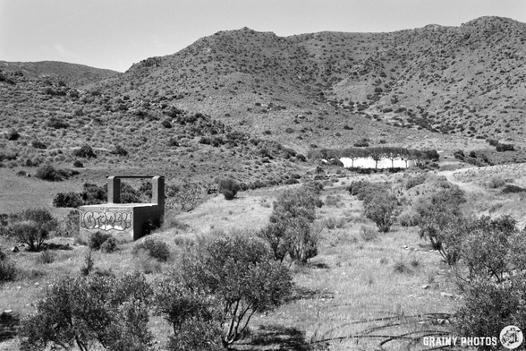 A desolate landscape featuring a barren field dotted with shrubs and a weathered concrete structure bearing graffiti. In the background, rolling hills rise under a clear sky, contributing to an atmosphere of abandonment and nature reclaiming its space.