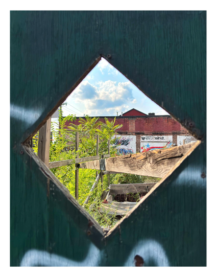 An empty city lot, seen through a cutout in a plywood barrier. The cutout is diamond-shaped, set in a green painted surface with some wood grain and streaks of white spraypaint. Through this we see a space overgrown with weeds as well as some makeshift scaffolding made of wooden planks and beams. In the background is a low brick building wall with some rectangular sections painted white and overlaid with graffiti. Above is a blue sky with puffy cumulus clouds.