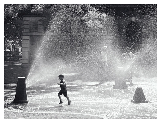 Black and white photo of a child running through the sunlit spray of a playground fountain. The child is shadowed in the foreground, contrasting with the bright spray of droplets. Through the mist we can make out some adults standing in the background, as well as some park trees and the small building for restrooms and park supplies.