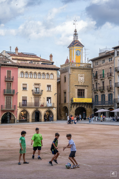 Color photo of a large dirt-covered plaza in a Catalonian town with four boys kicking around a football in the foreground and a clock tower rising above a yellow building in the background in the far corner of the plaza.