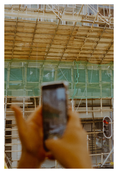 Color photography on the streets of Hong Kong. It is a blurry close-up of two hands holding up a smartphone. In the background is a multi-story building whose facade is covered with bamboo scaffolding and green safety nets typical of Hong Kong. On the right, a red pedestrian traffic light is lit. The bright photo is rendered in warm, slightly yellowish tones.