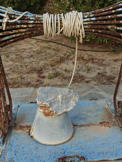 A photo of a covered well with a coiled white  rope wound around a rusted metal pulley, securing a bucket weighted down against the wind by a large stone.
