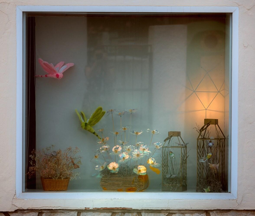Ein Schaufenster eines Blumenladens in einer Seitenstraße, mitttig ein Arrangement von Blumen aus Cellophanpapier, sie schimmern orange und tealfarben, zwei Plüschschmetterlinge in rosa und moosgrün. Abendstimmung, in der Spiegelung ein Müllcontainer, dessen Öffnung wie ein Auge aussieht, ich fotografiere an einer Haustür gelehnt.