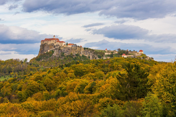 Ein herbstlicher Wald im Vordergrund. Dahinter ergebt sich ein Felsen mit einer Burg on top. Der Himmel ist bewölkt.