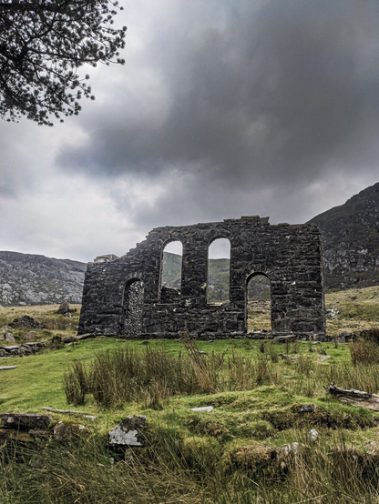 The ruins of a 19th-century chapel with arched windows and doors carved from dark stone sits in a green reedy field. In the background, rugged rocky mountains rise under a moody, overcast sky with dark clouds.