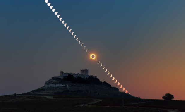 A photograph of a dimly lit castle silhouetted against the dusk sky. It's a combination of multiple exposures, with the Sun appearing as a sequence of discs crossing the sky diagonally above the castle. A progressively smaller fraction of the Sun is visible as the eclipse progresses. Right above the castle the eclipse reaches the total phase, with the Sun completely blocked by the Moon, revealing a fiery corona around it. The sequence continues towards the horizon, with the Sun progressively reappearing.