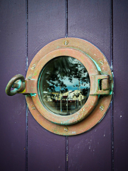 Close-up of a vintage round brass porthole window with a hinge and screw bolt lock. The weathered, oxidised metal is set into a deep purple paneled door. The reflection of townhouses, ship masts, and tree branches can be seen in the porthole glass.