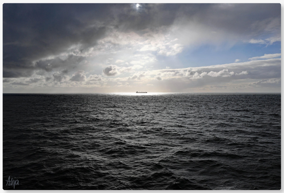 Auf der Fähre auf der Nordsee: Graue dicke Regenwolken schieben sich von links ins Bild, ein kleiner Rest blauer Himmel rechts. Hinter den Wolken die Sonne, die einen Lichtstrahl auf einen Tanker wirft und die graue See silbrig glänzen lässt.

On a ferry on the North Sea: thick, grey rain clouds drift into the frame from the left, with a small patch of blue sky visible on the right. Behind the clouds, the sun casts a beam of light onto a tanker and making the grey sea glisten silvery.