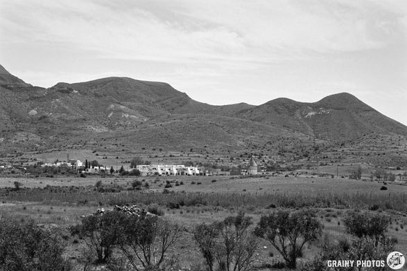 A black-and-white landscape featuring rolling hills and distant mountains with a small village nestled in a grassy valley, surrounded by scattered trees. The sky is partly cloudy, adding depth to the serene rural scene.