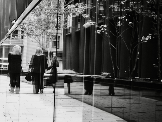 Three women office workers captured walking between two tall modern buildings lined with plate-glass walls, where their reflections are disrupted by the glass and surrounding marble so that only their lower legs can be seen as they pass a couple of tall trees.