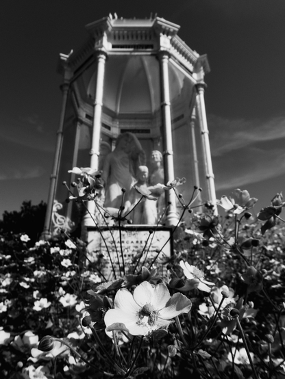 Flowers growing in a thick border around an ornate glass and stone memorial sculpture in Stirling. The white marble sculptures stand out against a clear blue sky.