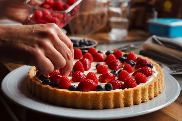 auf dem bild ist eine obsttorte mit cremiger füllung zu sehen, die mit frischen erdbeeren, himbeeren und blaubeeren belegt ist. eine hand dekoriert gerade die torte. sie steht auf einem weißen teller auf einem holztisch, während im hintergrund weitere früchte und küchenutensilien zu sehen sind.
