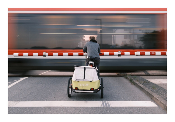 Ein Radfahrer mit einem Fahrradanhänger wartet vor einem geschlossenen Bahnübergang. Ein roter Zug fährt gerade vorbei. Der Zug ist durch seine Bewegung unscharf abgebildet.

A cyclist with a bike trailer is waiting in front of a closed railroad crossing. A red train is just passing by. The train appears blurred due to its movement.