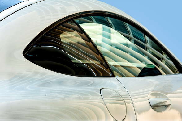 Deutsch: Das Bild zeigt eine Detailaufnahme der hinteren Seitenpartie eines weißen Autos. Im oberen Bereich dominiert ein dunkel getöntes Seitenfenster mit einer bogenförmigen Kontur, die nach hinten spitz zuläuft. Auf der weißen Karosserie sind im unteren Bereich die Naht einer runden bis leicht ovalen Tankklappe sowie am rechten Rand ein in Wagenfarbe lackierter Türgriff zu sehen. Über den Lack und die Fensterscheibe ziehen sich markante Spiegelungen, die geschwungene, helle architektonische Streben oder Lamellen vor einem blauen Himmel zeigen und sich der Wölbung des Fahrzeugs anpassen. English: The image shows a close-up of the rear side section of a white car. The upper area is dominated by a dark-tinted side window with a curved outline that tapers to a point towards the rear. On the white bodywork, the seam of a round to slightly oval fuel filler flap is visible in the lower section, along with a body-colored door handle on the right edge. Prominent reflections stretch across the paint and the window glass, showing curved, light-colored architectural struts or louvers against a blue sky, which contort to the curvature of the vehicle. 